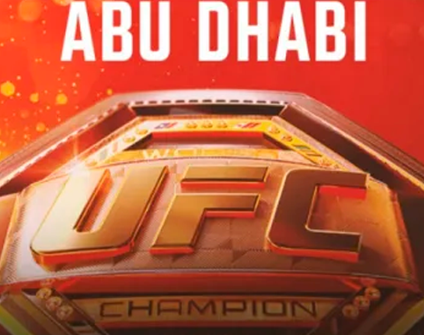 На изображении крупный план чемпионского пояса UFC с надписью Abu Dhabi на фоне. Пояс выполнен в золотисто-металлической цветовой гамме с надписью Champion внизу. Красный фон с искрящимися деталями